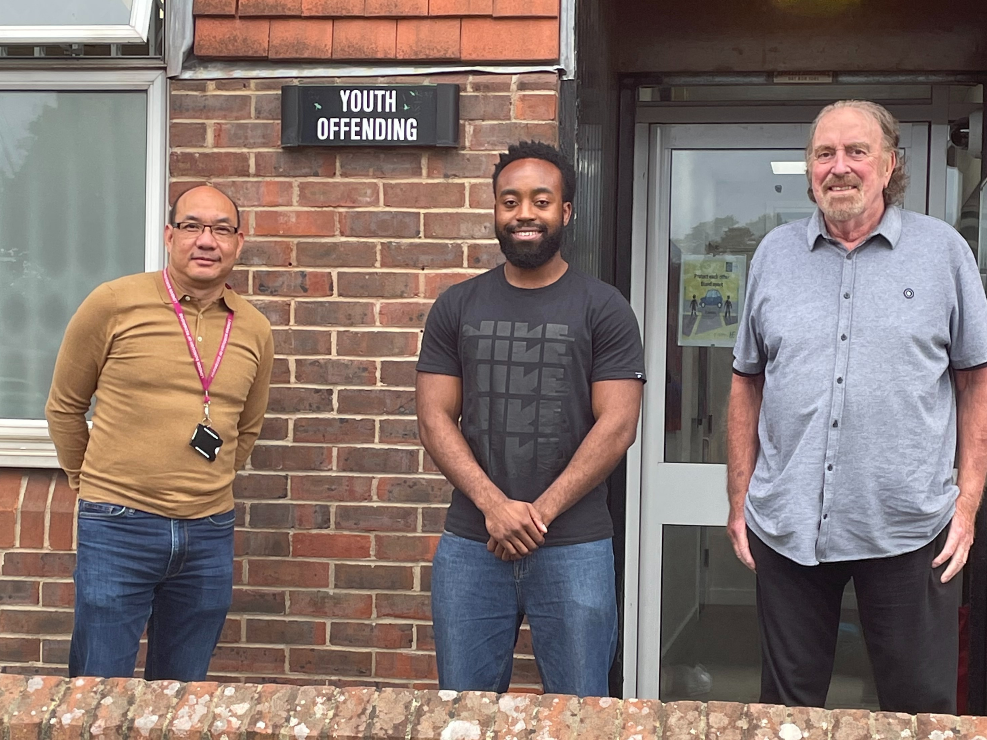 Three men face the camera stood outside a brick building